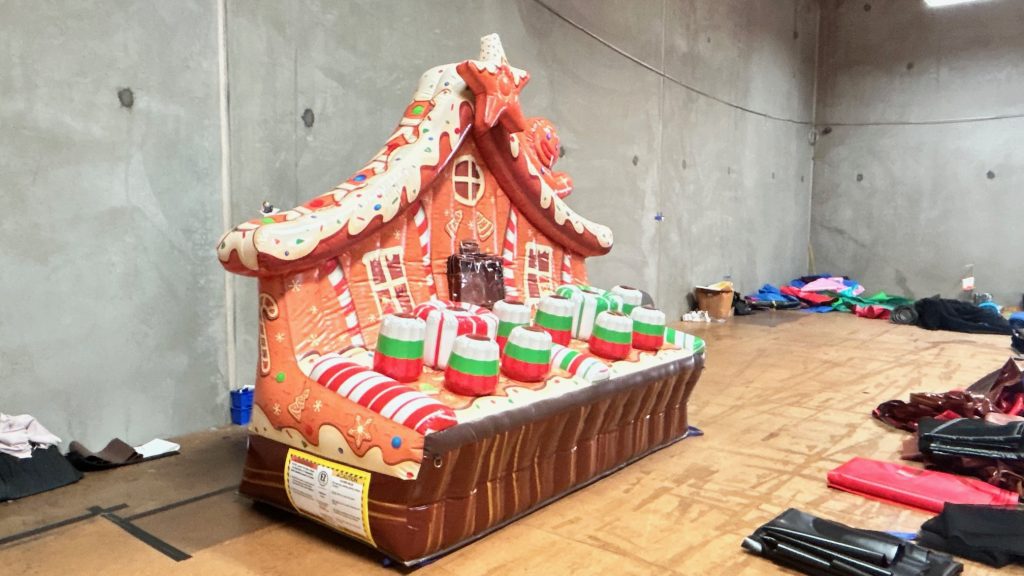 An inflatable gingerbread house with candy decorations sits in a large, empty indoor space, as if awaiting a Light Speed Gingerbread Crumble amidst concrete walls and scattered materials on the floor nearby. - SuperGames